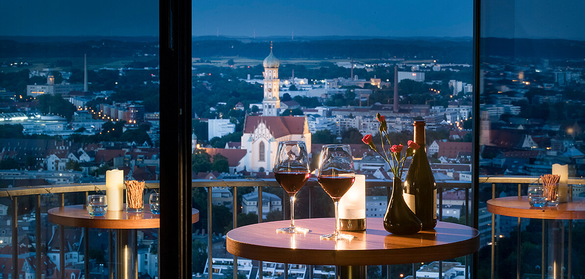 Zwei Gläser Rotwein stehen auf einem Tisch mit Blick auf eine nachts beleuchtete Stadt.